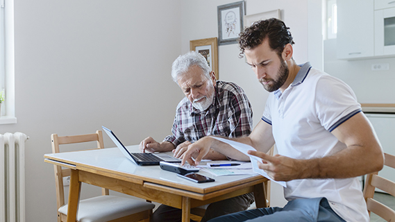 Son Doing Accounts Together With His Senior Father at Home, Planning New Purchase. Family Budget and Finances. Man Helping Senior Neighbor With Paperwork