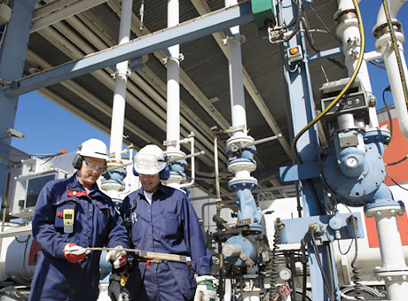 two workers in safety equipment reviewing equipment in front of large industrial pipes and equipment